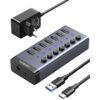 Na zdjęciu znajduje się hub USB marki UGREEN z siedmioma portami USB oznaczonymi jako "USB3.0" oraz przyciskami i niebieskimi diodami. Obok huba widoczny jest zasilacz z wtyczką oraz dwa kable USB, z których jeden ma końcówkę USB Typu C, a drugi USB Typu 