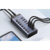 Na zdjęciu znajduje się hub UGREEN z siedmioma portami USB, z których cztery są zajęte przez podłączone kable. Na urządzeniu widoczne są przyciski oraz niebieskie diody, a na obudowie znajduje się logo "UGREEN".