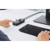 Na zdjęciu znajduje się hub USB z kilkoma podłączonymi urządzeniami, obok którego widoczna jest klawiatura oraz ręce osoby obsługującej te urządzenia. Hub jest podłączony do monitora, a na biurku nie ma innych widocznych elementów.