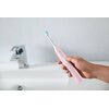 Różowa szczoteczka soniczna trzymana w dłoni, z widocznym napisem "Philips Sonicare" na uchwycie. W tle znajduje się biały zlew z metalową baterią.