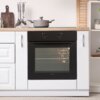 Czarny piekarnik elektryczny FINLUX FO-65E0N3F0BK jest wbudowany w białą kuchenną zabudowę, z widocznymi dwoma pokrętłami na panelu sterowania. Na blacie nad piekarnikiem stoi szary garnek z pokrywką, a obok znajdują się przybory kuchenne i deska do kroje