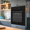 Czarny piekarnik elektryczny FINLUX FO-65W1E8F0BK jest wbudowany w szafkę kuchenną, z widocznym panelem sterowania z pokrętłami i wyświetlaczem cyfrowym. W tle znajduje się kuchnia z drewnianymi półkami i roślinami.