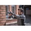 Na zdjęciu widoczna jest osoba używająca wiertarko-wkrętarki BOSCH Professional GSB 18V-90 C 06019K6100 Solo do wiercenia w ceglanej ścianie. Osoba ma na sobie ochronne okulary i zatyczki do uszu, a w tle widać budynek z oknami zabezpieczonymi kratami.