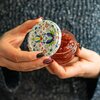 Osoba trzyma w dłoniach słoik z przetworami, a w drugiej ręce ozdobną zakrętkę z kolorowym, kwiatowym wzorem. Tło jest rozmyte, a paznokcie osoby są pomalowane na ciemne kolory.