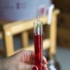 Na zdjęciu widoczny jest winomierz zanurzony w cylindrze z czerwonym płynem, trzymany w dłoni. Na winomierzu znajdują się oznaczenia liczbowo-kolorystyczne, a w tle rozmyty jest drewniany mebel.