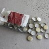 Na zdjęciu widoczna jest otwarta paczka kapsli do butelek marki BROWIN, z której część kapsli wysypała się na szarą, perforowaną powierzchnię. Na opakowaniu znajduje się logo BROWIN oraz napis "Kapsle do butelek 100 szt.".