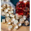 Na zdjęciu widoczne są złote zakrętki do butelek, rozłożone na drewnianej powierzchni, obok przezroczystej torby z większą ilością zakrętek. W tle znajdują się szklane butelki wypełnione ciemnoczerwonym płynem oraz szklanka z podobnym napojem.