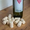 Na zdjęciu znajduje się butelka z etykietą przedstawiającą kiść winogron i napis "Wine", obok której leży kilka jasnych korków do butelek na drewnianej powierzchni.