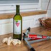 Na zdjęciu znajduje się zielona butelka z etykietą przedstawiającą kiść winogron i napis "Wine", obok której leży kilka korków do butelek oraz metalowe narzędzie z czerwonym elementem. W tle widoczny jest drewniany blat i biała ściana z płytkami.