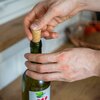 Na zdjęciu widoczne są dłonie wkładające korek do zielonej butelki z etykietą przedstawiającą liść i czerwone owoce. W tle znajduje się rozmyty fragment kuchni z drewnianym blatem.