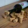 Na zdjęciu znajduje się zielona butelka wina z korkiem w szyjce, leżąca na drewnianej desce, wokół której rozłożone są korki do butelek. Etykieta na butelce przedstawia czerwone owoce i zielone liście.