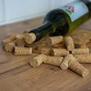 Na zdjęciu widoczna jest leżąca butelka z korkiem oraz rozrzucone wokół niej korki do butelek na drewnianej powierzchni. Etykieta na butelce przedstawia czerwone owoce i zielone liście.