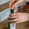 Na zdjęciu widoczne są ręce wkładające korek do zielonej butelki z etykietą przedstawiającą kiść winogron. W tle znajduje się rozmyty fragment kuchni z drewnianym blatem.