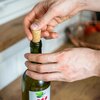 Na zdjęciu widoczne są dłonie wkładające korek do szyjki zielonej butelki z etykietą przedstawiającą liść i czerwone owoce. W tle znajduje się rozmyty fragment kuchni z koszem na owoce.
