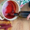 Na zdjęciu widać brązową butelkę zanurzaną w czerwonym wosku znajdującym się w ceramicznym naczyniu z roślinnym wzorem. Obok leży kilka czerwonych lasek wosku na drewnianej desce.
