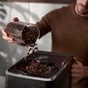 Szklanka z ziarnami kawy jest przechylona nad otworem ekspresu do kawy, a ziarna wsypują się do środka. Ekspres ma ciemną obudowę z widocznymi przyciskami i pokrętłami na górze.