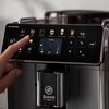 Na zdjęciu widoczny jest panel sterowania ekspresu do kawy z wyświetlaczem dotykowym, na którym znajdują się ikony różnych rodzajów kaw, takich jak Espresso, Coffee, Americano, Cappuccino, Latte Macchiato, Caffè Latte oraz More Drinks. Na dolnej części ek