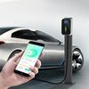 Na zdjęciu znajduje się ładowarka do samochodu elektrycznego zamontowana na słupku, obok której stoi nowoczesny samochód. W dłoni widoczny jest smartfon z aplikacją pokazującą parametry ładowania: "0.00 kW", "0.0 A", "230V", "Current Charging Energy", "To