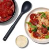 Na zdjęciu widoczna jest czarna łyżka obok talerza spaghetti z klopsikami w sosie pomidorowym, udekorowanego liśćmi bazylii i posypanego serem. Obok znajduje się miseczka z tartym serem oraz patelnia z klopsikami w sosie pomidorowym.