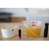 Na zdjęciu znajduje się miarka skośna OXO Good Grips 250 ml z przezroczystego plastiku z czarnym uchwytem, wypełniona płynem o jasnym kolorze, z widoczną czerwoną skalą pomiarową i logo "OXO". Obok niej stoi biały pojemnik z suchym produktem oznaczony ety