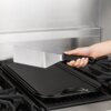 Na zdjęciu widoczna jest ręka trzymająca metalową szpatułkę z czarną rączką nad czarną patelnią grillową na kuchence gazowej. W tle znajduje się stalowa powierzchnia, prawdopodobnie część okapu kuchennego.