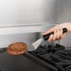 Na zdjęciu widoczna jest metalowa szpatułka z czarną rączką, trzymająca brązowy kotlet nad płytą grzewczą. W tle znajduje się stalowa powierzchnia kuchenna.