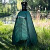 Zielony namiot turystyczny stoi na trawie w pobliżu wody, a na jego szczycie umieszczony jest czarny worek z napisem "NILS CAMP SHOWER NC1711". W tle widoczny jest zalesiony krajobraz z jeziorem.