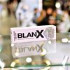 Pudełko pasty do zębów z napisem "BLANX Non-abrasive whitening toothpaste" oraz "WHITENING 100% NATURAL ARCTIC LICHENS" stoi na szklanej powierzchni, odbijając się w niej. Tło jest rozmyte, z widocznymi kolorowymi światłami i postaciami.