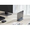 Na zdjęciu znajduje się biurko z monitorem, klawiaturą i myszką, a obok stoi tablet umieszczony w stojaku do laptopa MOZOS H1. W tle widoczne są zasłony, a na biurku leżą rozrzucone zdjęcia.