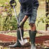 Osoba pracuje w ogrodzie, używając szpadla prostego marki FISKARS Ergonomic 1067512 do przekopywania ziemi. W tle widoczne są podwyższone grządki z roślinami oraz ogrodzenie.