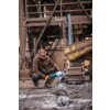 Mężczyzna w brązowej bluzie roboczej i okularach ochronnych używa szlifierki kątowej w przemysłowym otoczeniu, otoczony metalowymi konstrukcjami i resztkami materiałów na ziemi. Na zdjęciu widoczne są elementy maszyn i konstrukcji przemysłowych.