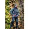 Na zdjęciu znajduje się osoba ubrana w zielono-czarną odzież outdoorową, stojąca na tle skalistego, porośniętego mchem zbocza. Osoba ma na sobie czapkę i plecak, a na kurtce widoczne jest logo.