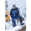 Na zdjęciu widoczna jest osoba ubrana w niebiesko-czarną kurtkę i spodnie robocze, trzymająca szlifierkę kątową na tle zaśnieżonego terenu z metalowymi elementami i roślinnością. Osoba ma na sobie szare rękawiczki i czapkę, a w tle znajduje się biały kont