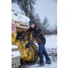 Na zdjęciu widać osobę w kurtce roboczej LAHTI PRO L4093806 (rozmiar 3XL) stojącą obok żółtej maszyny budowlanej z widocznym logo "KOMATSU", na tle zaśnieżonego terenu i drzew. Osoba ma na sobie czapkę i rękawice, a na maszynie widoczny jest śnieg.