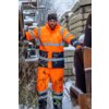 Mężczyzna w pomarańczowym, odblaskowym stroju roboczym stoi na tle zaśnieżonego terenu z betonowymi blokami i metalowymi elementami. Na ziemi leży śnieg, a w tle widać zarośla i drzewa.