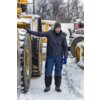 Mężczyzna w zimowej kurtce i niebieskich spodniach roboczych stoi na zaśnieżonym placu obok dużej maszyny budowlanej pokrytej śniegiem. W tle widoczne są inne maszyny oraz drzewa.
