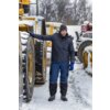 Mężczyzna w zimowej kurtce i niebieskich spodniach roboczych stoi na zaśnieżonym terenie obok dużej maszyny budowlanej. W tle widoczne są inne maszyny pokryte śniegiem.