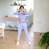Na zdjęciu znajduje się osoba ćwicząca z fioletowym hula hop w jasnym, nowoczesnym wnętrzu z drewnianą podłogą i rośliną w doniczce. W tle widoczna jest kuchnia z półkami, na których stoją różne przedmioty i rośliny.