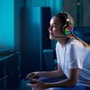 Na zdjęciu znajduje się osoba siedząca na kanapie, trzymająca kontroler do gier i nosząca słuchawki z mikrofonem, które mają podświetlenie RGB. W tle widoczny jest ekran telewizora oraz elementy wyposażenia wnętrza.