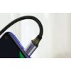 Na zdjęciu widoczny jest czarny, pleciony kabel USB-C podłączony do niebieskiego urządzenia, z metalową końcówką, na której znajduje się napis "UGREEN". Kabel jest lekko zgięty, a tło jest rozmyte i jasne.