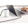 Na zdjęciu widać laptopa podłączonego do źródła zasilania za pomocą czarnego kabla USB-C, który jest wpięty do białej listwy zasilającej. Obok laptopa znajduje się ręka osoby piszącej na klawiaturze.