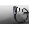 Na zdjęciu widoczny jest czarny, pleciony kabel USB-C z kątową wtyczką, na której znajduje się napis "UGREEN". Tło jest szare z delikatnym wzorem.
