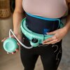Osoba trzyma zielony hula hop z przymocowanym ciężarkiem, który jest owinięty wokół talii. Na czarnym pasie widoczny jest niebieski napis "ONE FITNESS".