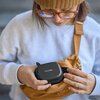Osoba trzyma czarne etui z napisem "WIRELESS GO II" i wkłada je do brązowej torby na ramię. Etui ma teksturowaną powierzchnię i zamek błyskawiczny.