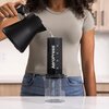 Na zdjęciu widoczny jest zaparzacz AeroPress XL, do którego wlewana jest woda z czarnego czajnika. Produkt stoi na przezroczystym pojemniku, a na jego powierzchni znajduje się napis "AeroPress".
