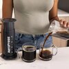 Na zdjęciu widoczny jest zaparzacz AeroPress XL obok dwóch szklanek z kawą, z których jedna jest napełniana z przezroczystego dzbanka. Na zaparzaczu znajduje się napis "AeroPress".
