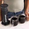 Na zdjęciu widoczny jest zaparzacz do kawy z napisem "AeroPress" oraz dwie szklanki z czarną kawą na blacie. Obok zaparzacza znajduje się osoba w szarej koszulce, trzymająca jedną z szklanek.