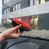 Na zdjęciu widać rękę trzymającą gąbkę z dyfuzorem na płyn, skierowaną w stronę szyby samochodowej, za którą znajduje się nowoczesny budynek z dużymi oknami. Gąbka ma czerwony materiał na wierzchu i białą dyszę do rozpylania płynu.