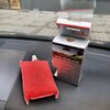 Na zdjęciu znajduje się czerwona gąbka z dyfuzorem oraz otwarte pudełko z napisem "NIGRIN" i informacjami o produkcie, w tym wymiarami "40 ml - 8 x 12 x 4 cm". Pudełko stoi na desce rozdzielczej samochodu, a w tle widać fragmenty parkingu.