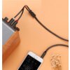 Na zdjęciu widoczny jest czarny kabel audio z wtyczką Jack 3.5 mm podłączony do urządzenia mobilnego oraz dwoma wtykami RCA podłączonymi do drewnianego głośnika. Na ekranie urządzenia mobilnego wyświetlana jest aplikacja do odtwarzania muzyki, a obok leżą